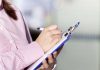 a supervisor in a pink jacket makes marks on a clipboard with a pen.